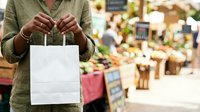 Unrecognizable person in olive shirt holding blank white paper bag at farmers market with fresh produce and vendors in soft focus background.