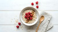 Creamy oatmeal porridge topped with fresh raspberries and granola in a ceramic bowl, wooden spoon on linen napkin, white wood background.