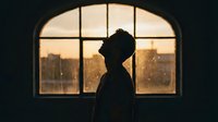 Silhouette of contemplative man standing by arched window at sunset with urban cityscape view. Moody atmospheric lighting creates dramatic contrast.