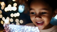 Young African American girl with curly hair smiling joyfully while looking at glowing lights, creating magical bokeh effect in warm evening atmosphere.