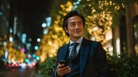 Professional Asian businessman in elegant suit using smartphone on illuminated city street at night with warm bokeh lights creating festive atmosphere.