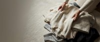 Woman in beige outfit sitting on textured floor organizing folded neutral-toned clothing and accessories in minimalist setting for lifestyle content.