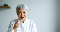 Senior Asian woman with short white hair wearing white chef's coat, smiling enthusiastically while holding wooden spoon, isolated on white background for culinary, cooking, and food service design projects.