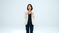 Young woman with dark hair and fair complexion wearing cream blazer and navy pants, gesturing with both hands in encouraging or welcoming pose against white background for business and lifestyle content.