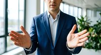 Professional businessman in navy suit gesturing with open hands during presentation in modern office with large windows and plants in background.