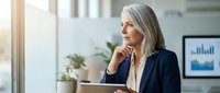 Mature woman with gray hair and Caucasian ethnicity wearing professional navy blazer holding tablet in modern office environment with natural light and plants.