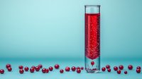 Tall glass test tube filled with sparkling red liquid surrounded by fresh cranberries on turquoise background, modern minimalist drink photography for beverage industry.