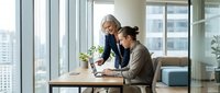 Senior businesswoman mentoring young female colleague at laptop in modern office workspace with city view windows for corporate training concepts.