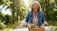 Happy mature Caucasian woman riding bicycle with flower basket on sunny tree-lined path, expressing joy and freedom in outdoor leisure activity.