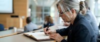 Professional mature businesswoman with glasses writing notes in modern office environment during productive work session.