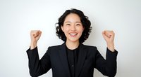 Young Asian businesswoman with short curly hair wearing black blazer celebrating success with raised fists and cheerful expression against white background.
