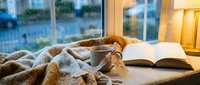 Cozy rainy day scene with warm knit blanket, tea cup, and open book on windowsill, soft lamp light, hygge home comfort atmosphere.