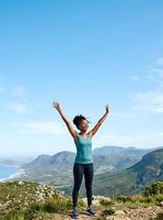 Young woman with dark hair and medium skin tone wearing athletic apparel, standing on mountain summit with arms raised in celebration, overlooking scenic valley landscape with coastal views for fitness, wellness, and outdoor lifestyle content.