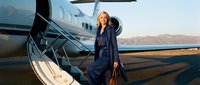 Confident blonde businesswoman in navy suit standing by private jet stairs at airport runway during golden hour for luxury travel and corporate success.