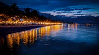 Coastal beach at twilight with golden streetlights reflecting on calm water, mountains silhouetted against blue dusk sky, perfect for travel and tourism designs.