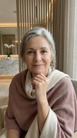 Mature woman with gray hair wearing mauve and cream layered sweater, hand near face, smiling at camera in modern luxury interior with warm lighting and vertical blinds.