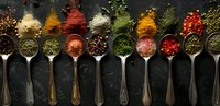 Colorful spices and herbs in vintage silver spoons arranged in row on dark background, overhead view. Aromatic seasoning collection with peppercorns and dried powders.