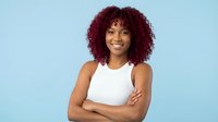 Confident young African American woman with red curly hair, arms crossed, smiling against light blue background, studio portrait.