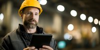 Bearded construction engineer in yellow hard hat using digital tablet at industrial site with bokeh lights in background.