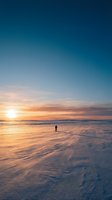 Solitary figure wading in shallow ocean waters during golden hour sunset, with warm amber and orange sky reflecting on wet sand and calm waves for travel and lifestyle content.