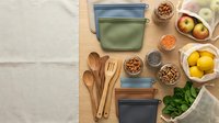 Flat lay overhead view of reusable food storage containers with wooden utensils, fresh produce, and dried ingredients on natural linen background for meal prep and kitchen organization.