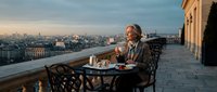 Mature Caucasian businessman enjoying coffee on elegant rooftop terrace overlooking historic European cityscape at golden hour sunset for luxury lifestyle content.