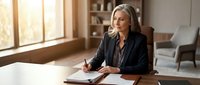 Professional mature Caucasian businesswoman in black blazer writing notes at wooden desk in modern office with natural sunlight streaming through windows.