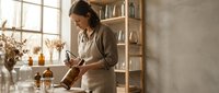 Young Asian woman potter shaping clay on wheel in bright ceramic studio workspace with natural light and wooden shelves for handmade pottery creation.