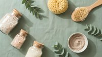 Spa and skincare products arranged flat lay with natural sea salt in glass jars, wooden bath brush, pumice stone, cream jar, and green fern leaves on sage background for wellness and beauty.