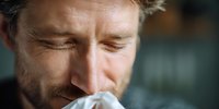 Close-up of middle-aged Caucasian man with stubble blowing nose into tissue, showing signs of cold or seasonal allergies against blurred background.