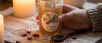 Herbal tea in glass jar with label held by hand near candles and dried botanicals on wooden surface for wellness and relaxation.
