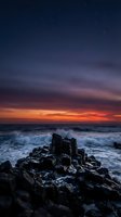 Dramatic coastal sunset with dark rocky outcrops in foreground, turbulent ocean waves, and vibrant orange-red sky transitioning to deep blue. Ideal for travel, nature, and environmental design projects.