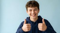 Young Caucasian teenage boy with curly brown hair smiling broadly while giving double thumbs up gesture, wearing navy blue hoodie against light background.