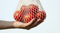Hand holding a reusable mesh net bag full of fresh red tomatoes on white background, eco-friendly grocery shopping concept.