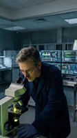 Middle-aged Caucasian male scientist in lab coat examining samples through microscope in modern research laboratory with computer monitors and equipment.