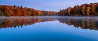Tranquil autumn lake with vibrant fall foliage reflected in calm water during misty morning twilight creating peaceful natural landscape scene.