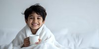 Young child wrapped in white blanket holding mug with warm drink, smiling happily against light background.
