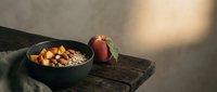 Rustic breakfast scene with black bowl of oatmeal topped with nuts and dried fruit, fresh apple, and wooden spoon on aged wooden surface with natural sunlight.