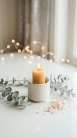 Cozy home spa setup with burning candle in ceramic holder, eucalyptus branches and pink salt crystals on white surface with warm bokeh lights background.