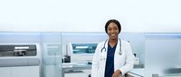 Professional African American female doctor in white coat with stethoscope standing confidently in modern hospital setting with medical equipment background.