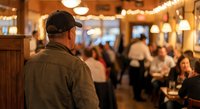 Male server in casual uniform working at busy restaurant with warm lighting and dining customers in background creating hospitality atmosphere.