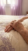 Elderly hands resting on white bedding with purple floral embroidery in bright bedroom with lavender curtains and natural sunlight streaming through window.