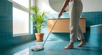 Young woman mopping turquoise tiled bathroom floor with modern wooden vanity and tropical plant creating clean fresh home environment for hygiene maintenance.