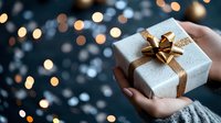 White gift box with golden bow held in female hands against blurred Christmas lights background creating magical holiday atmosphere and bokeh effect.