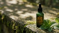 Natural herbal essential oil bottle with dropper on moss-covered stone surface surrounded by fresh green mint leaves in dappled sunlight for wellness.