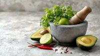 Fresh ingredients for guacamole preparation with stone mortar, avocado halves, lime, cilantro and chili peppers on concrete background.
