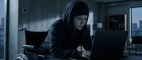 Young male hacker wearing dark hoodie working on laptop in dimly lit office space at night, engaged in cybercrime or data theft activity.