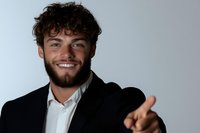 Young professional man with curly dark hair and beard wearing black suit and white shirt points and smiles confidently against light background.