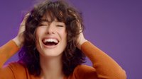 Young woman with curly hair laughing joyfully against purple background, wearing orange sweater, expressing genuine happiness and positive emotions.