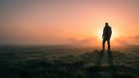 Silhouetted hiker standing in misty meadow at sunrise, watching golden light break through fog over rolling landscape and grassland terrain.